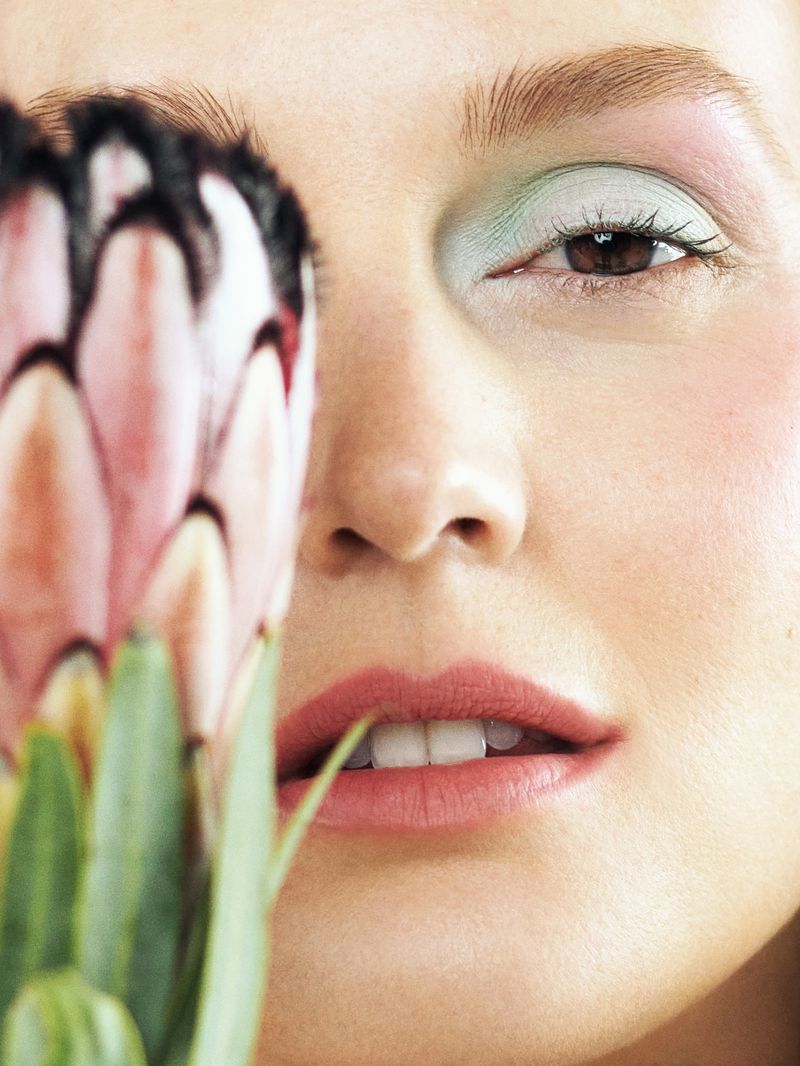 Close-up portrait of a woman with mint eyeshadow and pink blush, holding a protea flower, showcasing beauty and cosmetic artistry.