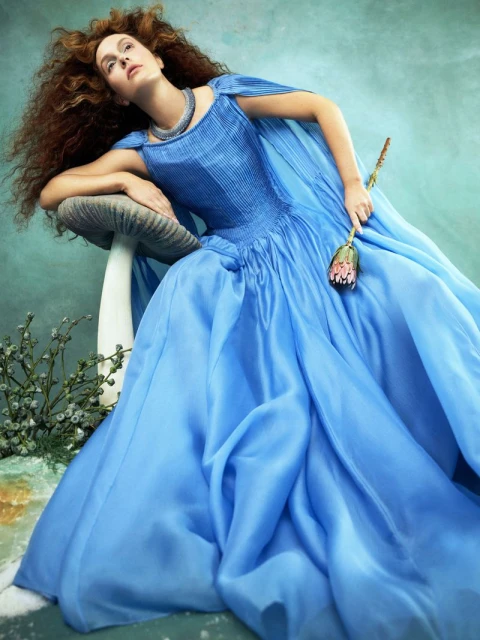 Woman in a blue gown poses next to a mushroom prop, holding a flower. She has red curly hair and wears a silver necklace.