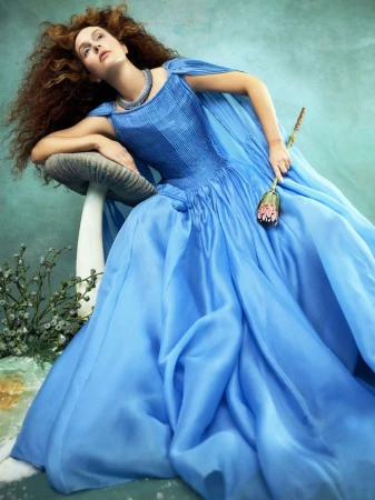 Woman in a blue gown poses next to a mushroom prop, holding a flower. She has red curly hair and wears a silver necklace.