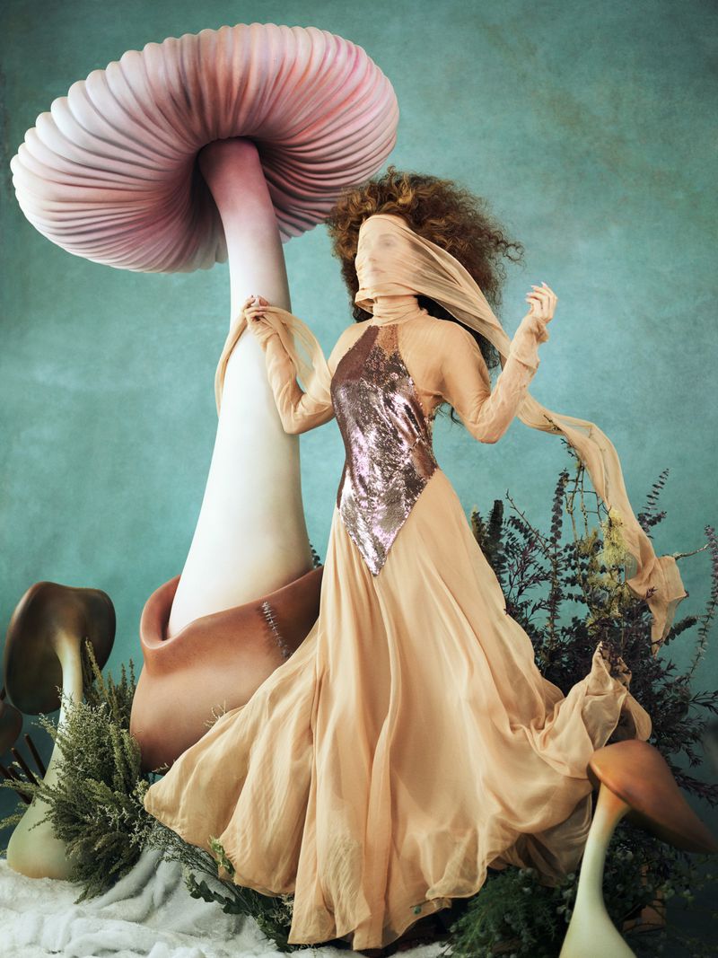 A person in a gown poses near large mushroom sculptures in a fantastical set, in a fashion-style photo with teal and beige tones.