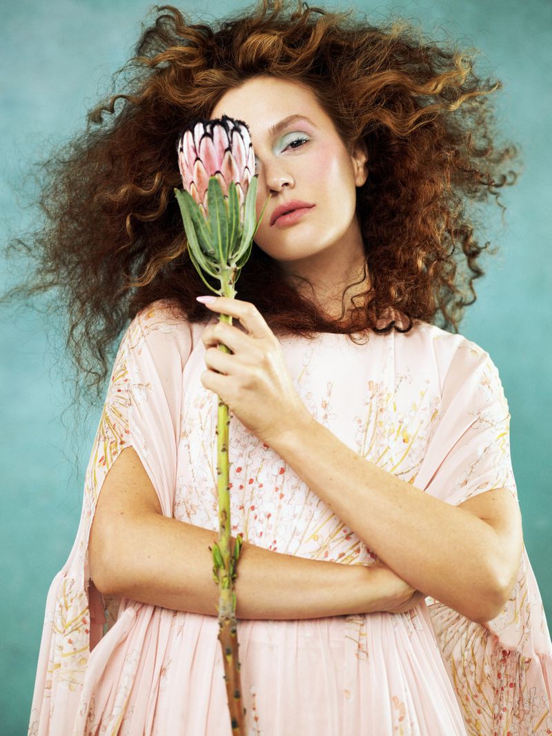 A young adult woman with curly hair poses indoors in a light pink dress while holding a large protea flower against a teal backdrop.