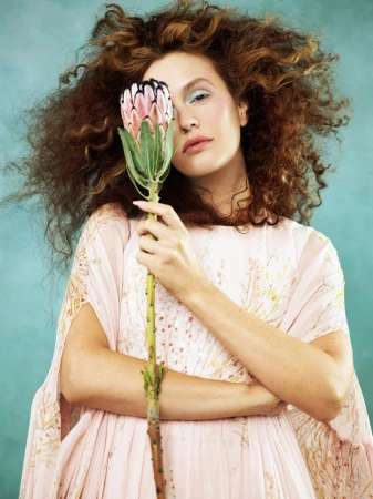 A young adult woman with curly hair poses indoors in a light pink dress while holding a large protea flower against a teal backdrop.