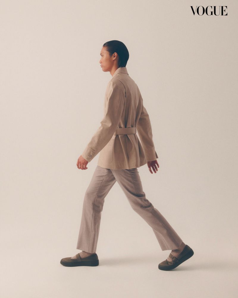 An adult female is captured in a studio shot, side profile, walking against a neutral backdrop. She wears a belted blazer, trousers, and loafers.