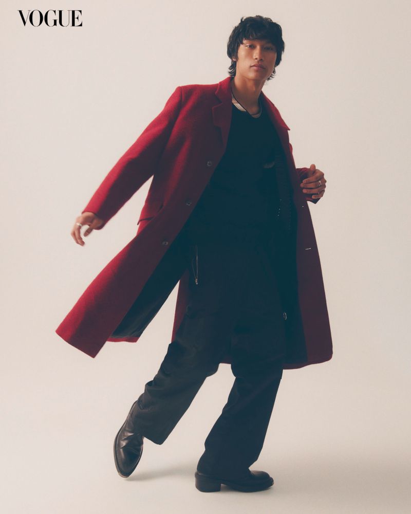 Fashion editorial shot of an adult male with medium dark hair wearing a red overcoat, black shirt and pants, posing in a photo studio.