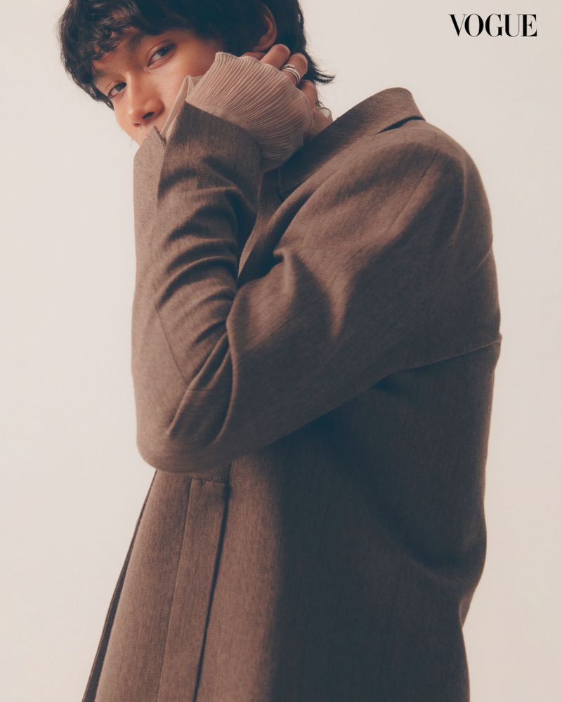 Fashion editorial shot of a young adult with dark, wavy hair in a brown coat and tan turtleneck sweater against a neutral backdrop.