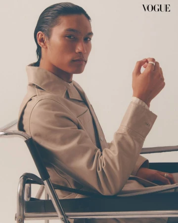 Portrait of an Asian man in a beige trench coat sitting in a silver wheelchair against an off-white background, fashion editorial.