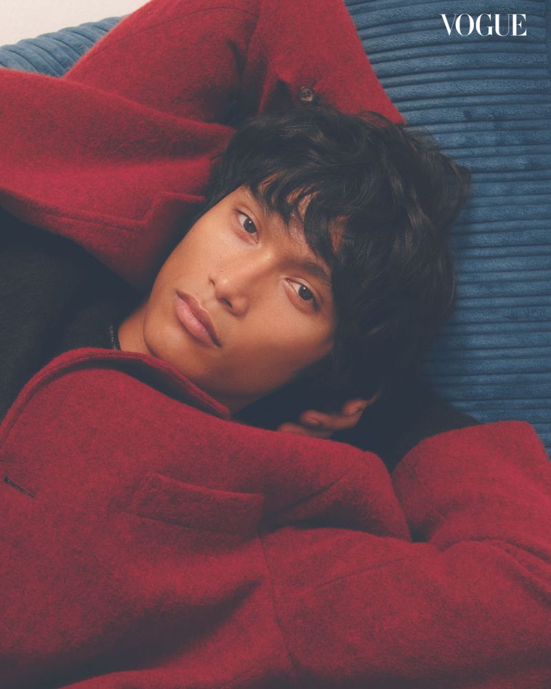Close-up studio portrait of a person with dark curly hair, wearing a red jacket, against a blue corduroy cushion and Vogue logo.