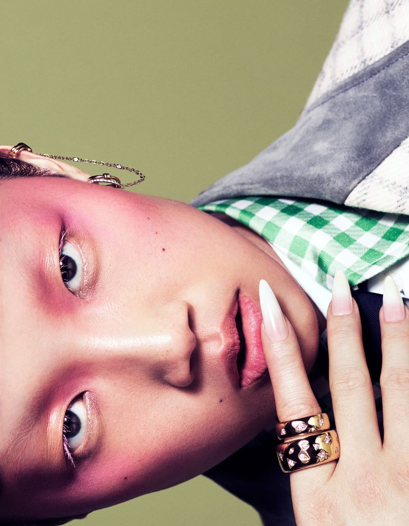 Close-up portrait of a model with dramatic pink makeup and extra-long white fingernails, wearing statement rings, against a green background.