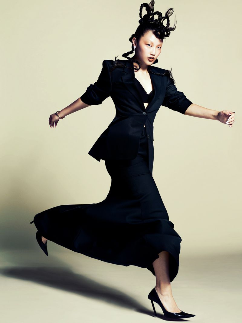 Portrait of an asian woman with braided hair posing in black blazer, skirt, and heels. She is in motion in a studio setting.
