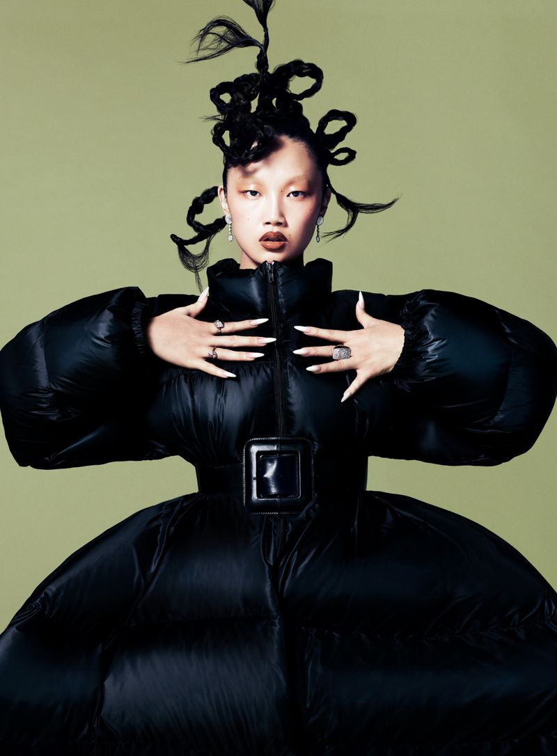 Close-up of an Asian female model wearing a black puffer jacket with exaggerated sleeves and a unique braided hairstyle against a green backdrop.