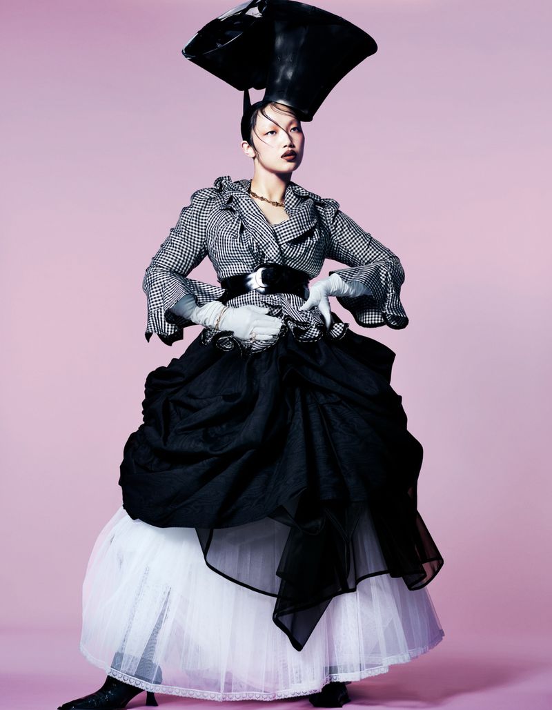 Asian woman in avant-garde fashion featuring a structured houndstooth jacket, voluminous black skirt, and a unique black headpiece.
