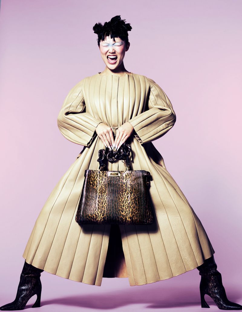 A person poses in a beige coat and black boots while holding a patterned purse against a lilac background in a studio setting.