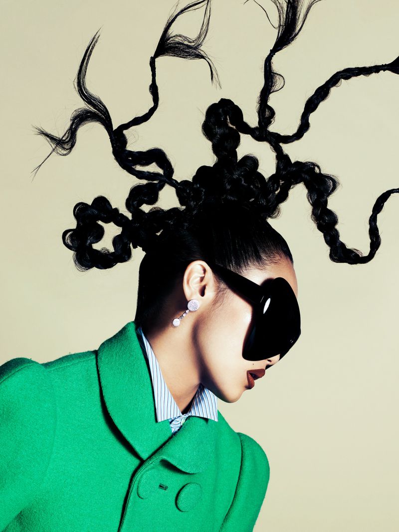 Woman in green coat and oversized sunglasses with elaborate black updo against cream background in eye-level studio portrait.