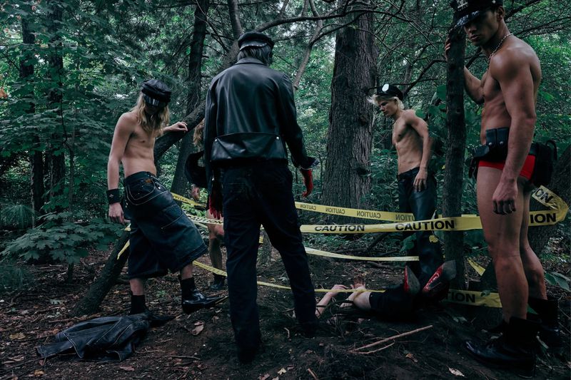 Group of bare-chested men in leather caps and fetish wear stand in a wooded area with yellow CAUTION tape wrapped around them.