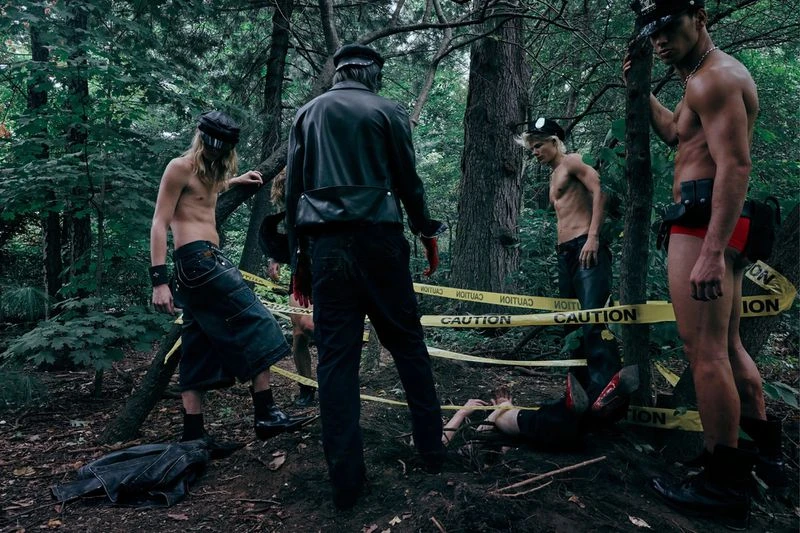 Group of bare-chested men in leather caps and fetish wear stand in a wooded area with yellow CAUTION tape wrapped around them.