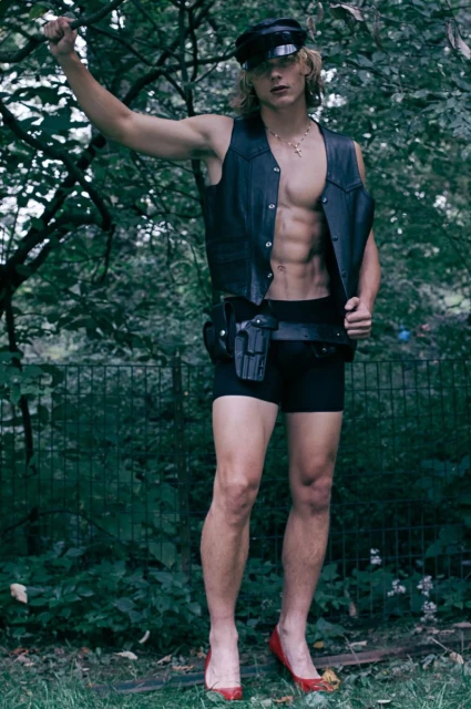 A slim, muscular man poses outdoors wearing a police cap, leather vest, and black shorts with red high heels in a natural setting.