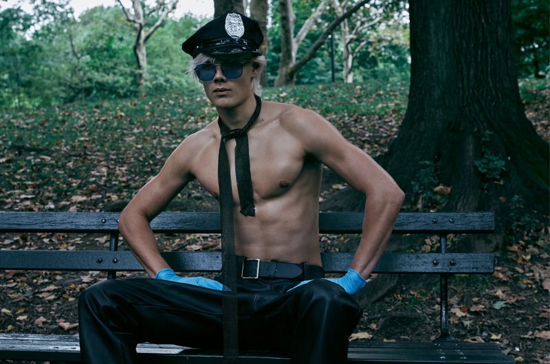 Shirtless man in police hat and blue gloves sits on park bench, wearing leather pants and tie, trees in background.