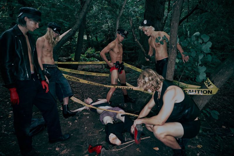 A group of scantily clad men and a prone woman are surrounded by yellow caution tape in a forest setting with dark and dramatic lighting.