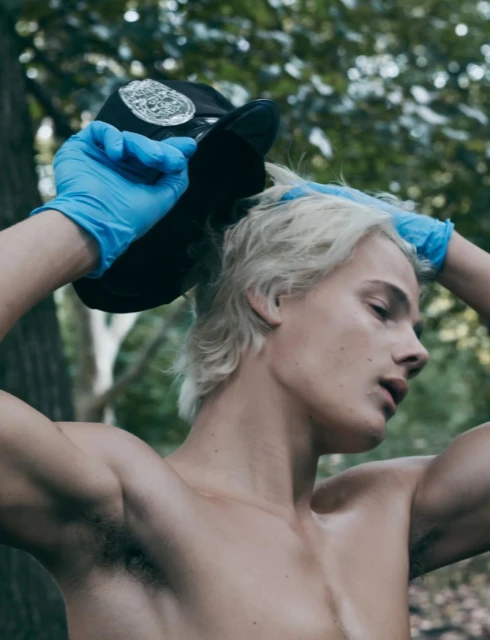 Shirtless man with blonde hair holds a cap in hands with blue gloves outdoors in a wooded environment with natural lighting.