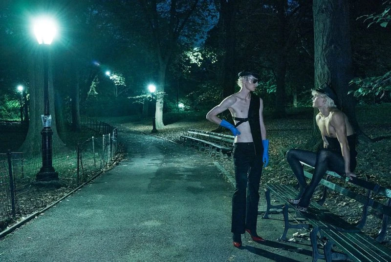 Two fair-skinned individuals pose in a nighttime park setting; one stands with blue gloves and red heels while the other sits on a bench.