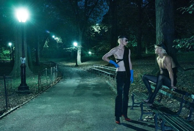Two fair-skinned individuals pose in a nighttime park setting; one stands with blue gloves and red heels while the other sits on a bench.