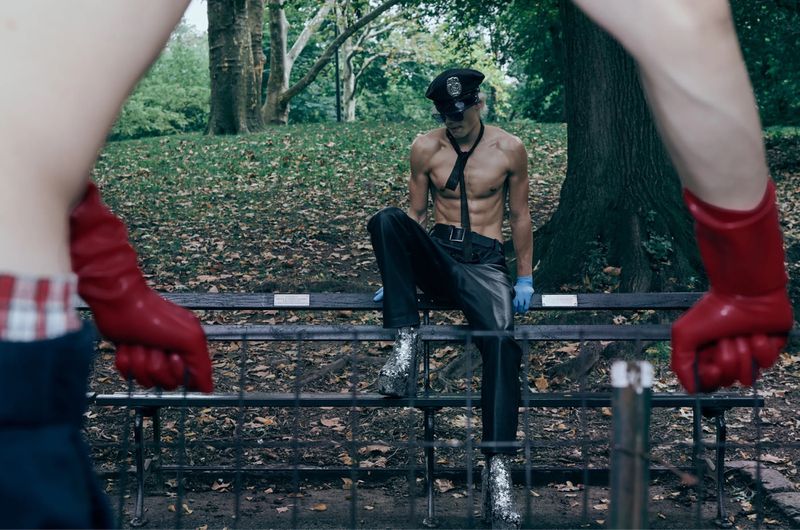 An adult male in a police hat and silver boots sits on a bench in a park, with another figure with red gloves in the foreground.