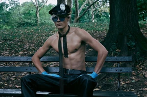 Shirtless male model wearing a police officer cap, sunglasses, black trousers, and blue gloves sitting on a park bench
