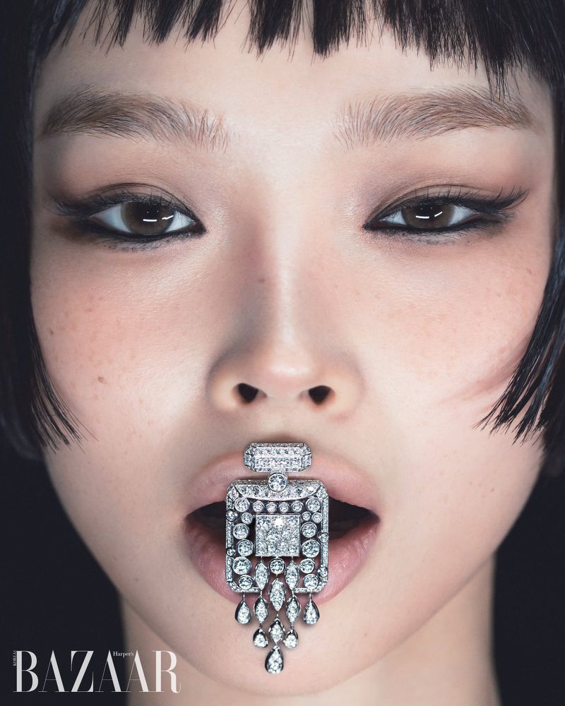 Close-up portrait of a model with dark eye makeup and fringe, holding a diamond jewelery pendant in her mouth against a black background.