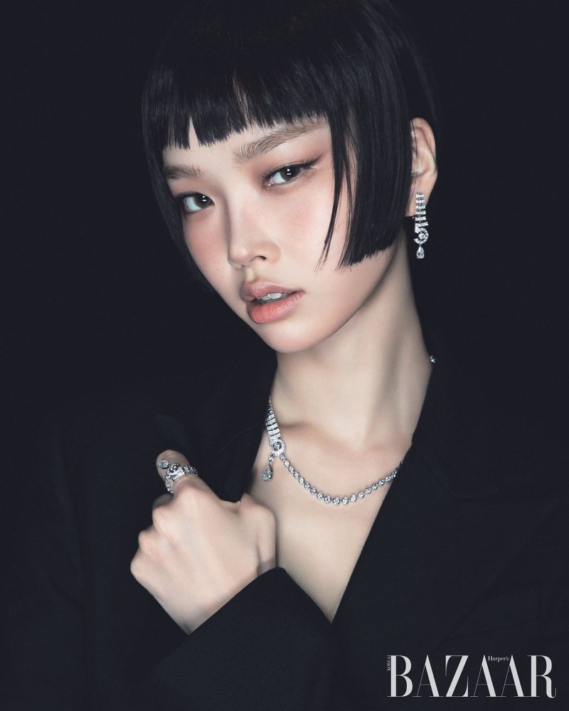 Close-up portrait of an adult female with short black hair and jewellery, wearing a black blazer against a dark background.