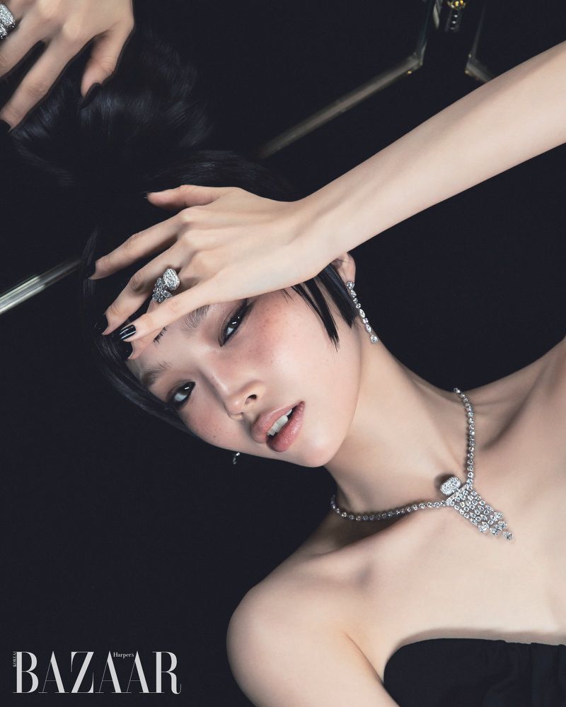 Portrait of a woman with dark hair and light skin, wearing a diamond necklace and ring, in a beauty and fashion shot against black.