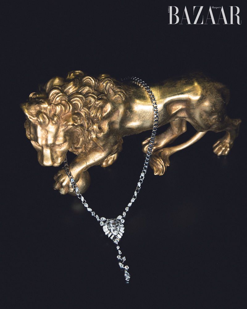 A gold lion sculpture supports a diamond necklace against a black backdrop, jewelry advertisement with a high-fashion magazine logo.