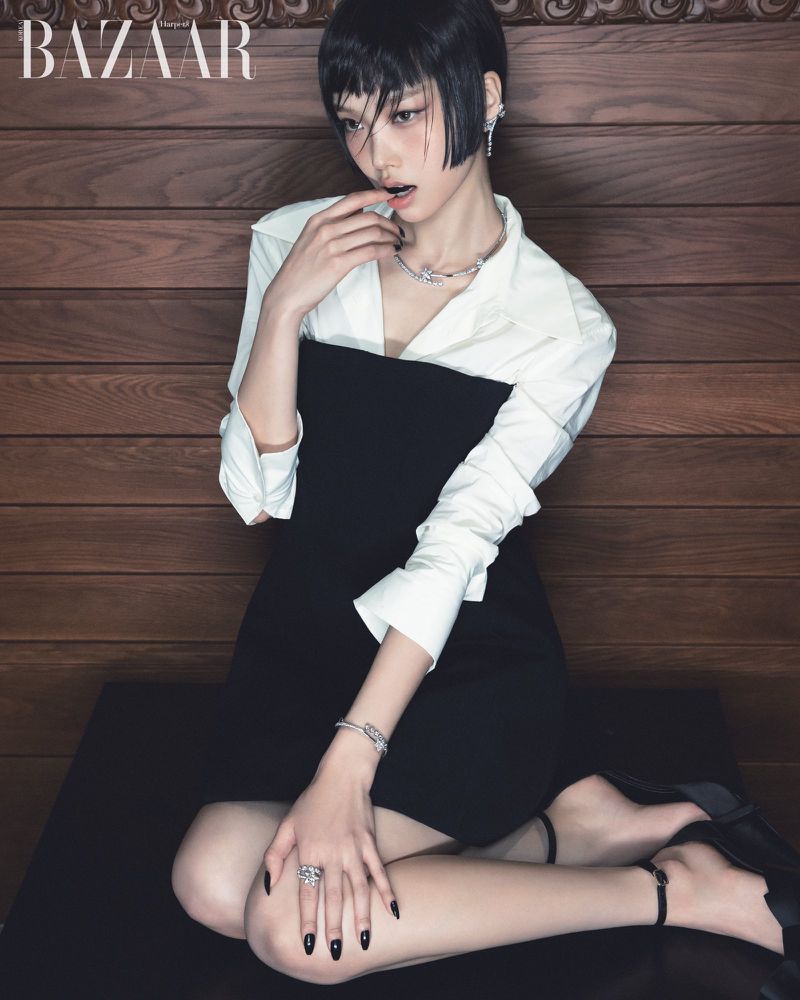 Young Asian woman in a black dress, jewelry, and white shirt poses indoors with a wood wall background for a fashion editorial.