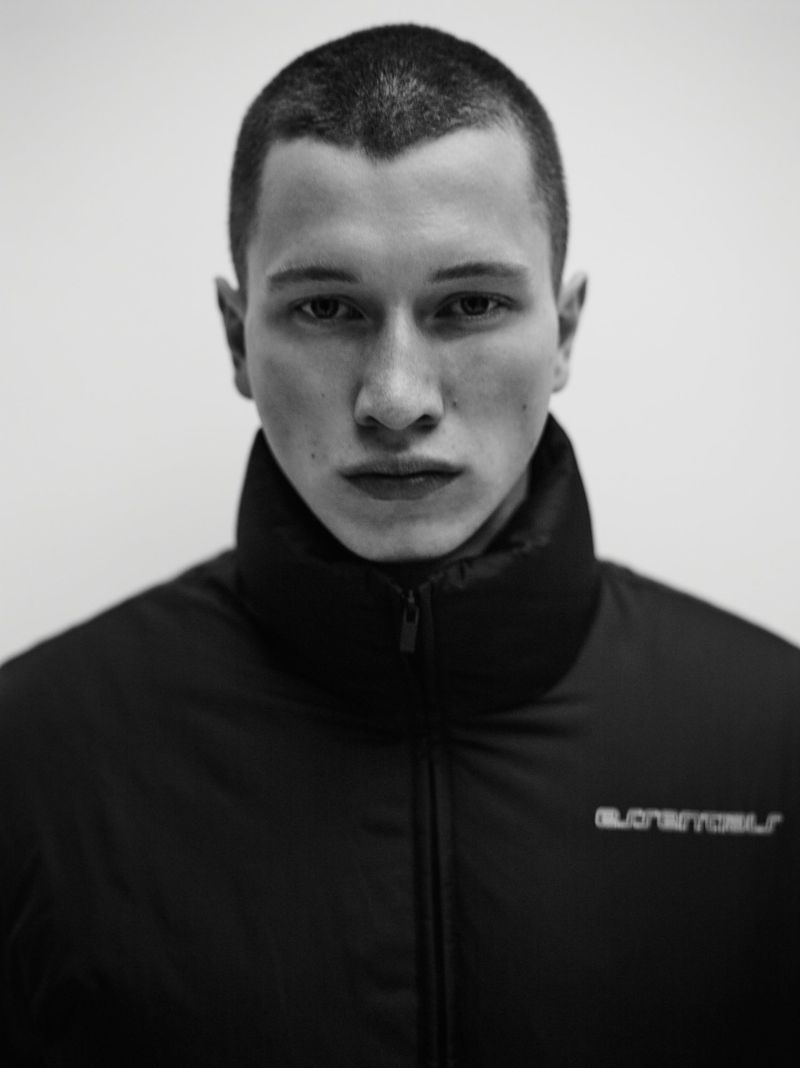 Monochrome portrait of a short-haired adult male in a high-collared black puffer jacket, set against a plain white backdrop.
