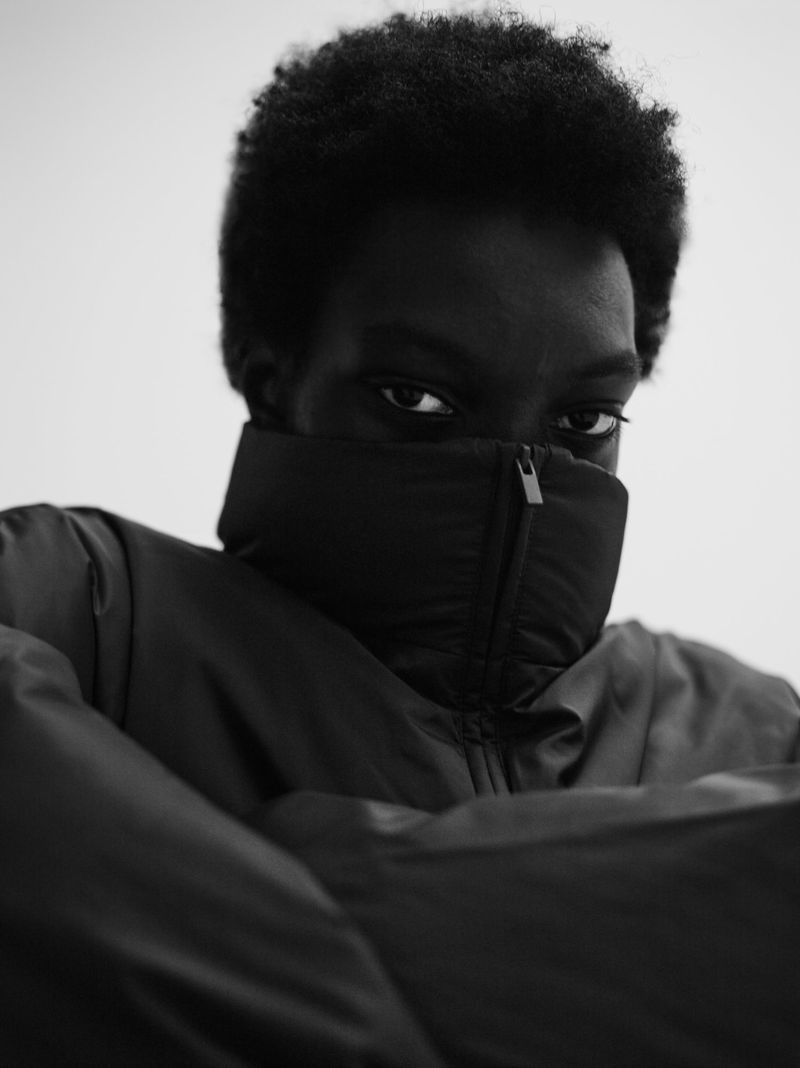 Black and white portrait of an African American adult with dark curly hair, wearing a black puffer jacket pulled up to cover the mouth.