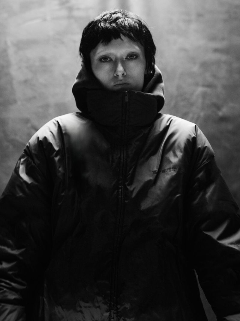 Monochrome portrait of a person wearing a puffy hooded jacket, set against a mottled grey backdrop in a studio-style setup.