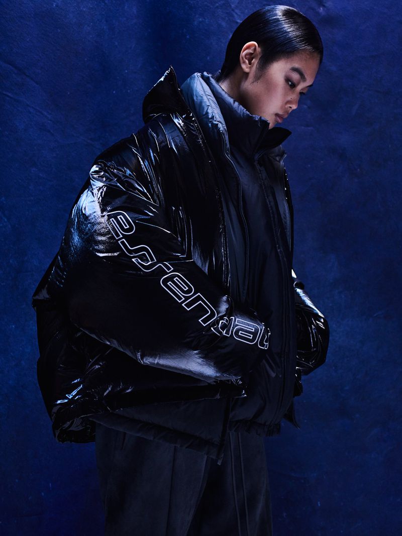 Woman in a shiny black puffer jacket with white logo, set against a dark blue backdrop with subtle textures and studio lighting.