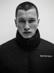 Close-up monochrome portrait of a young man wearing a high-collared black zip jacket against a plain light background
