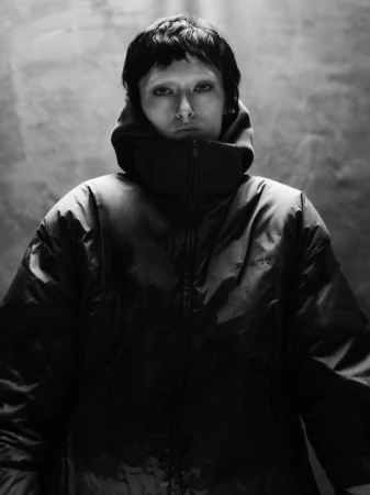 Monochrome portrait of a person wearing a zipped puffer jacket with a high collar against a textured background.