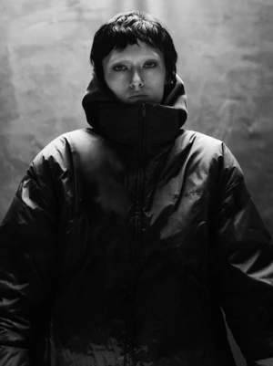 Monochrome portrait of a person wearing a zipped puffer jacket with a high collar against a textured background.