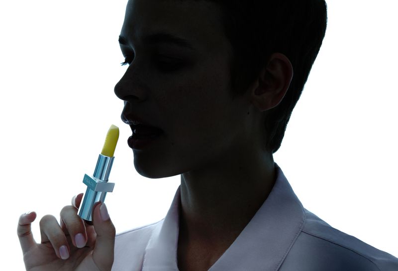 Woman applying yellow lipstick in a beauty close-up with a bright white background showcasing the product and the model's features.