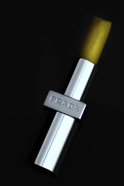 Close-up studio shot of a yellow and silver Prada lipstick tube. The background is solid black, and the lipstick is the primary focus.