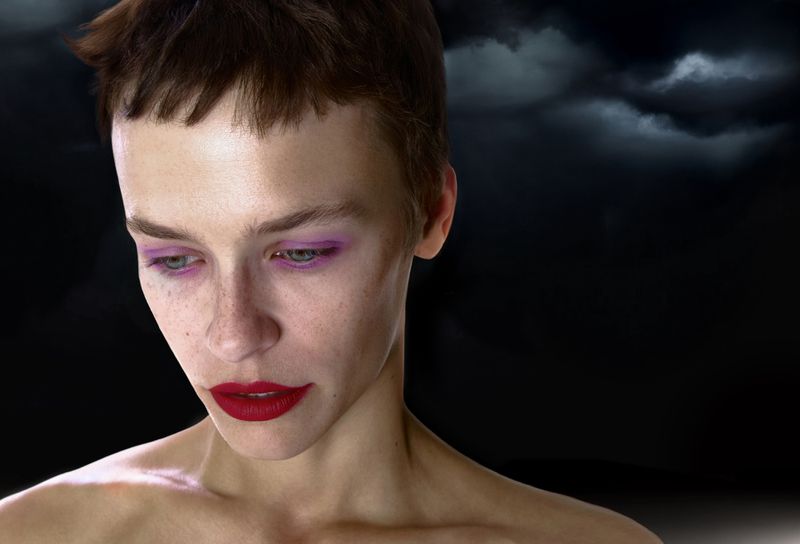 Close-up portrait of a model with short brown hair, purple eye shadow, and bright red lipstick against a dramatic dark cloudy sky