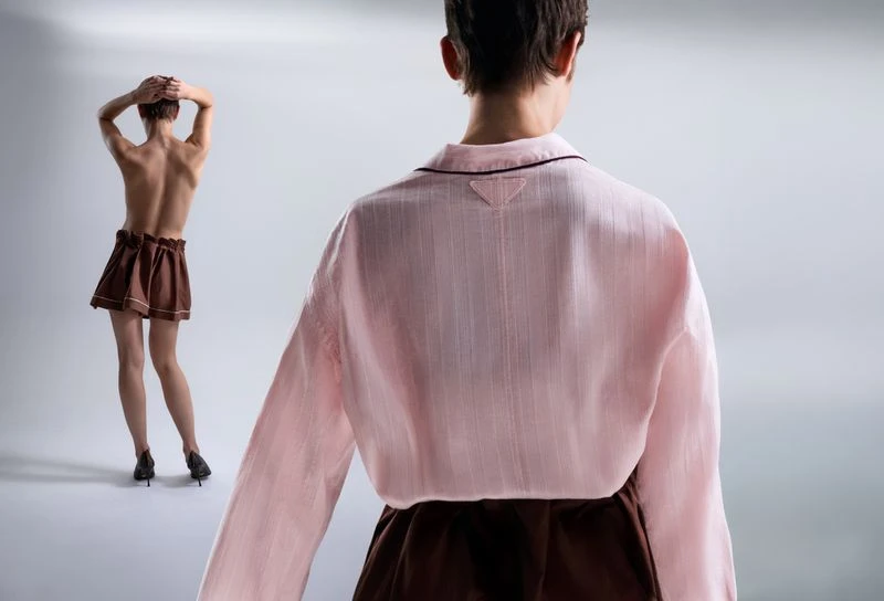 Two back-facing women in studio lighting. One woman in a pink top and brown skirt is close to the camera; the other is further away.