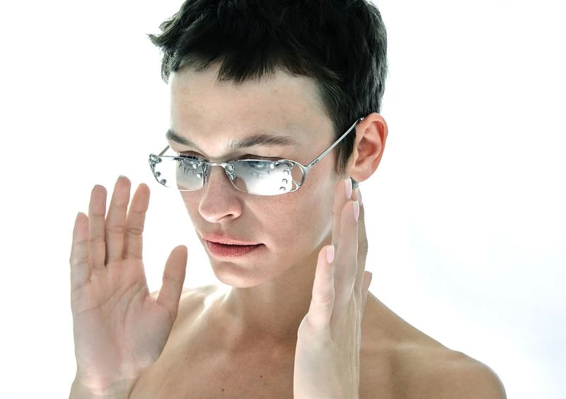 Adult woman posing indoors with silver-rimmed sunglasses, light skin, hands near face, and a white background. Beauty, fashion, eyewear.
