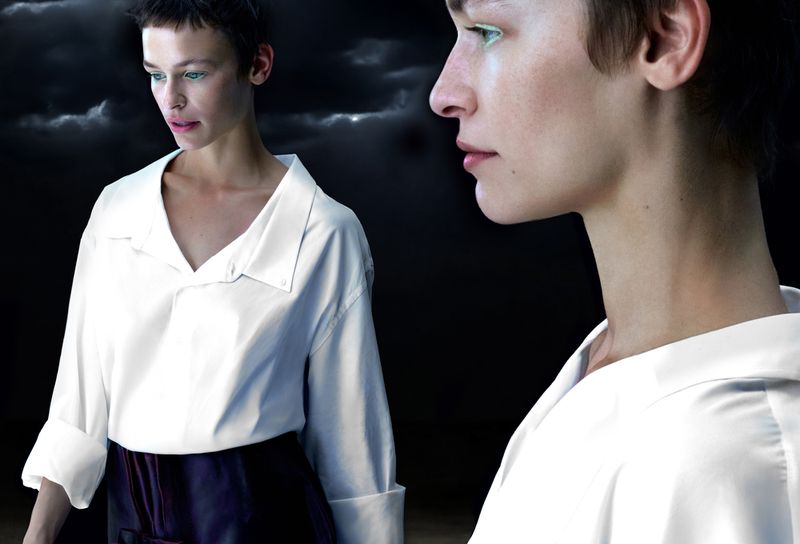 Fashion shot of a fair-skinned person in a white shirt and dark bottoms against a stormy sky background; studio lighting and dramatic mood.