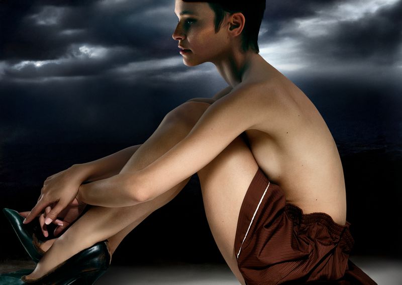 Adult female poses indoors with bare torso, brown shorts, and green high heels in a seated pose against stormy sky background.