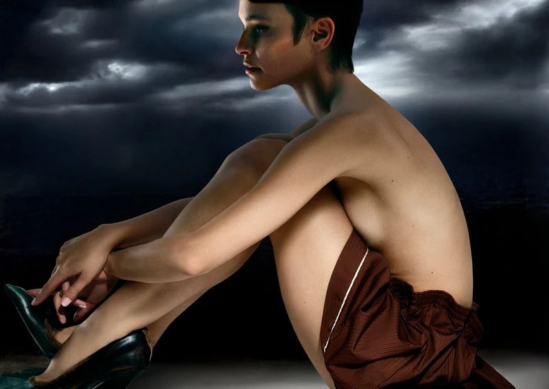 Adult female poses indoors with bare torso, brown shorts, and green high heels in a seated pose against stormy sky background.