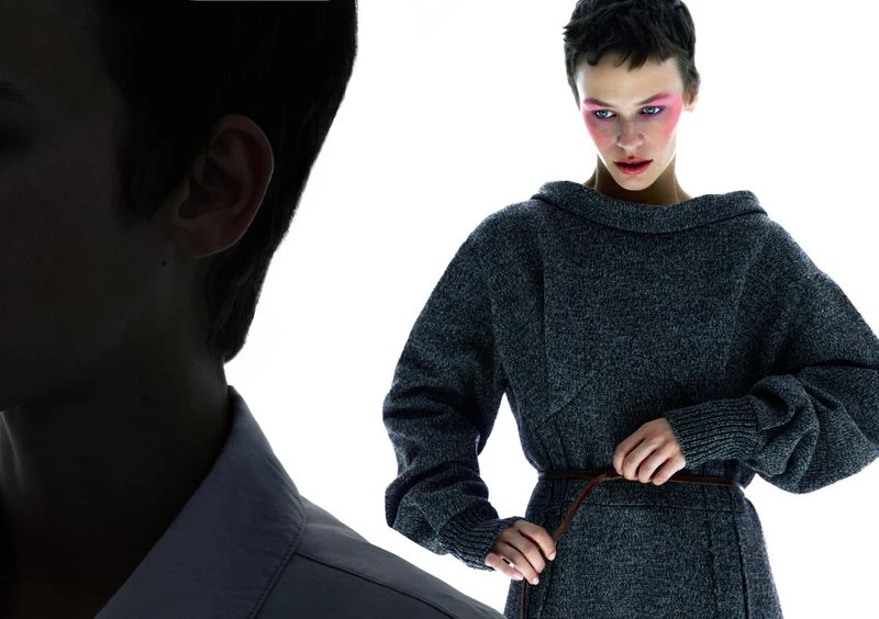 A woman with a short haircut wearing a gray dress cinched with a brown belt, styled with pink makeup, poses in a fashion shot.