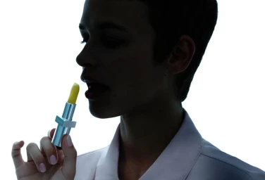 Close-up silhouette of a person applying yellow lipstick from a metallic case against a bright white background.