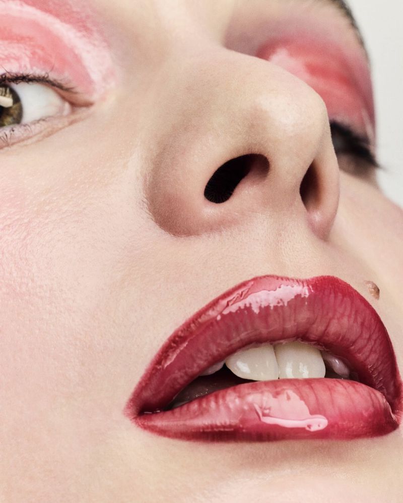 Close-up of a woman's face with glossy red lips and red eyeshadow in a beauty shot against a white background in portrait orientation.